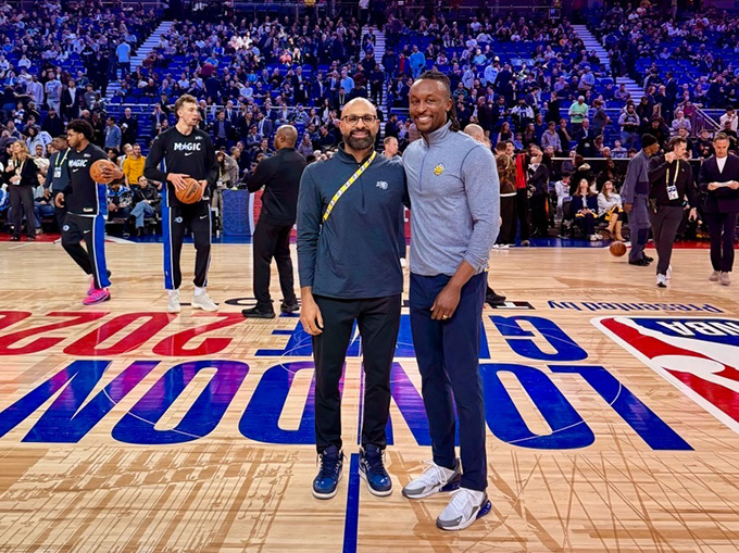 Jarred Boyd stands beside another man on a basketball court with team members warming up behind them and fans in the stands