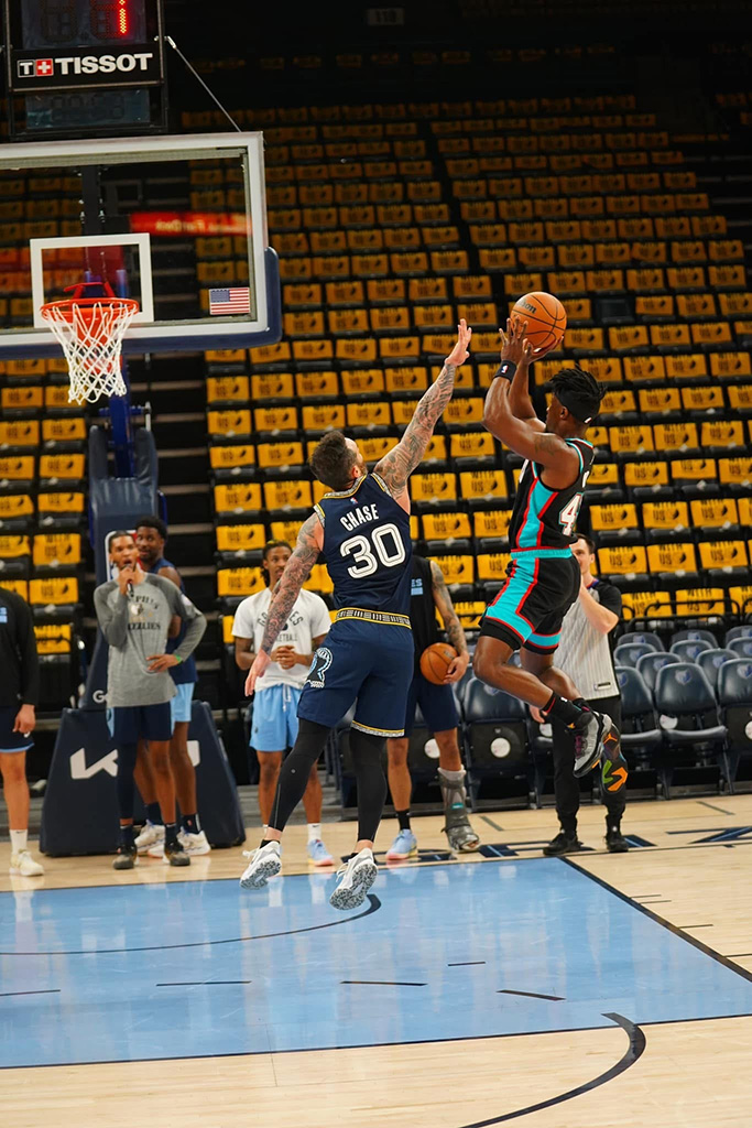A basketball player in uniform attempts a shot while another player on the opposite team raises his arm to block; several onlookers watch from the sidelines in an empty arena